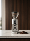 A silver electric coffee grinder with a clear hopper and digital display sits on a white marble countertop, with roasted coffee beans placed beside it against a dark wood-paneled background.
